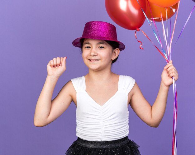 Feliz joven caucásica con gorro de fiesta violeta sosteniendo globos de helio y manteniendo el puño aislado en la pared púrpura con espacio de copia