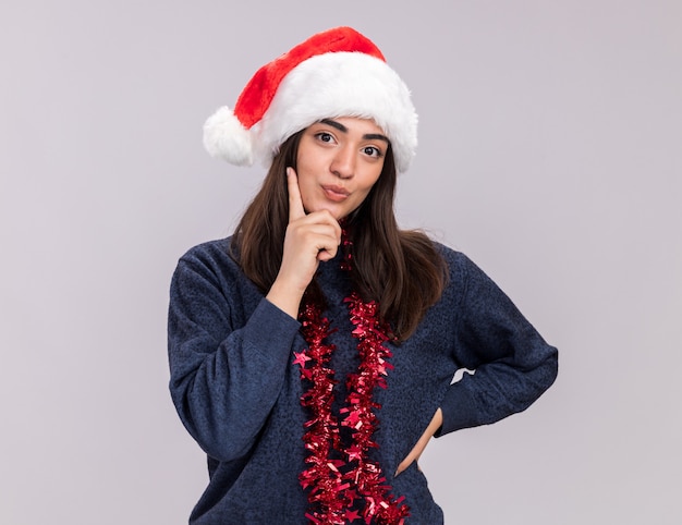 Feliz joven caucásica con gorro de Papá Noel y guirnalda alrededor del cuello pone el dedo en la barbilla y mira