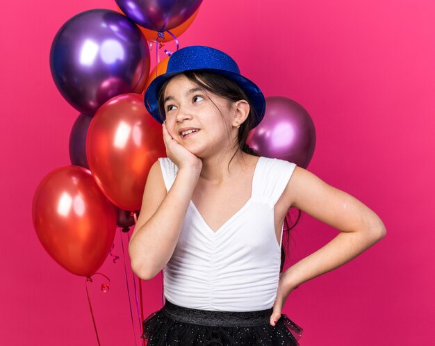 Feliz joven caucásica con sombrero de fiesta azul poniendo la mano en la cara y mirando al lado de pie frente a globos de helio aislados en la pared rosa con espacio de copia