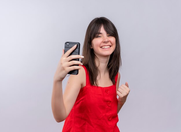 Feliz joven caucásica sosteniendo el teléfono sosteniendo el puño y mirando a la cámara sobre fondo blanco aislado