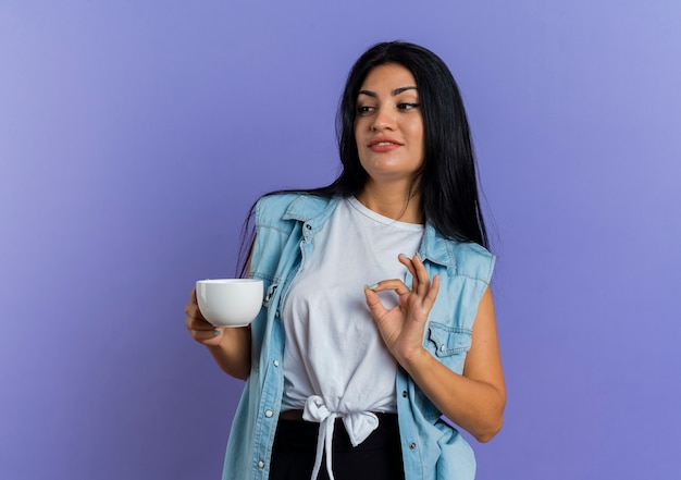 Feliz joven caucásica sostiene la taza y mira al lado aislado sobre fondo púrpura con espacio de copia