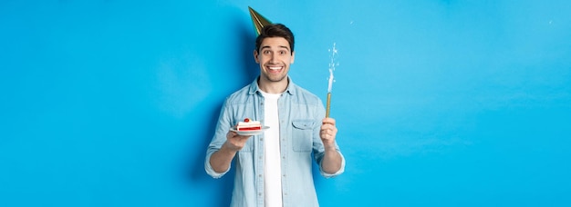 Foto gratuita feliz joven celebrando un cumpleaños con gorro de fiesta sosteniendo un pastel de cumpleaños y sonriendo de pie sobre una b azul