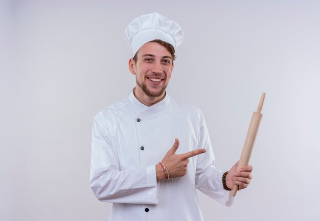 Un feliz joven chef barbudo hombre vestido con uniforme de cocina blanco y sombrero apuntando al rodillo mientras mira en una pared blanca