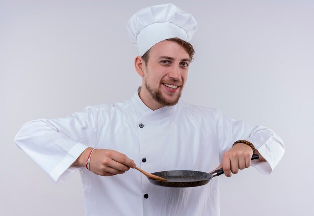 Un feliz joven chef barbudo hombre vestido con uniforme de cocina blanco y sombrero sosteniendo una cuchara de madera en una sartén mientras mira en una pared blanca
