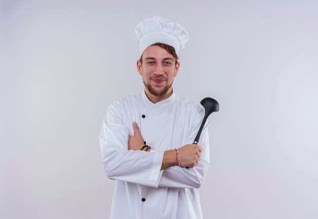 Un feliz joven chef barbudo hombre vestido con uniforme de cocina blanco y sombrero sosteniendo cucharón negro mientras mira en una pared blanca