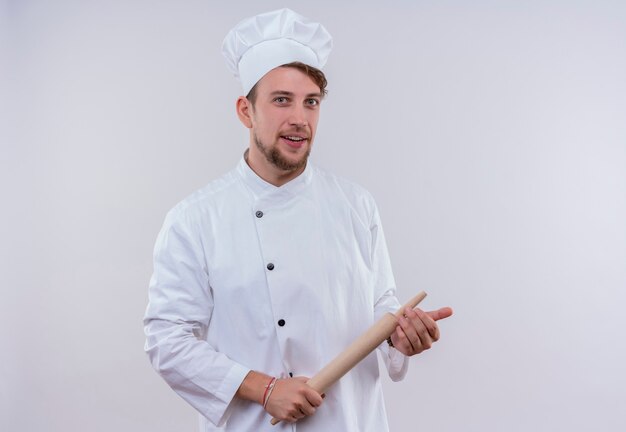 Un feliz joven chef barbudo hombre vestido con uniforme de cocina blanco y sombrero sosteniendo un rodillo mientras mira en una pared blanca