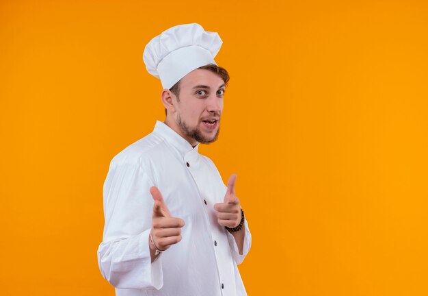 Un feliz joven chef barbudo con uniforme blanco apuntando con el dedo índice sobre una pared naranja