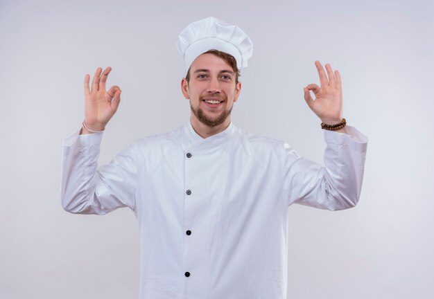 Un feliz joven chef barbudo en uniforme blanco mostrando gesto ok con las dos manos sobre una pared blanca