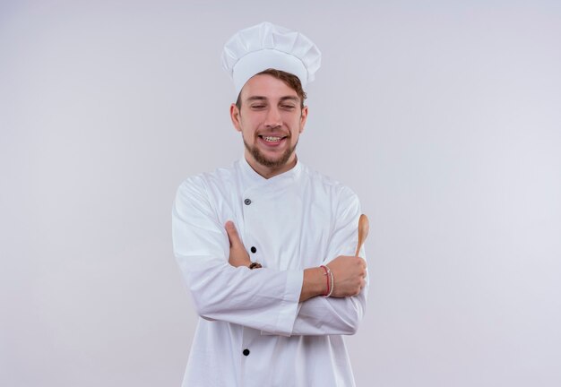 Un feliz joven chef barbudo con uniforme de cocina blanco y sombrero sosteniendo una cuchara de madera con los ojos cerrados mientras está de pie sobre una pared blanca