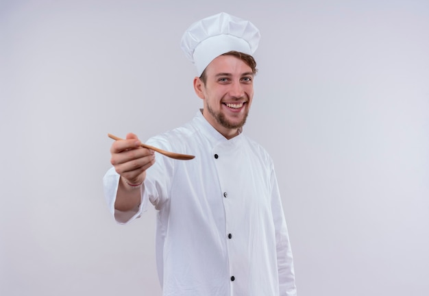 Un feliz joven chef barbudo vestido con uniforme de cocina blanco y un sombrero que invita a degustar con una cuchara de madera en la mano mientras mira una pared blanca