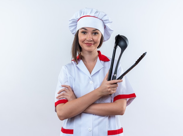 Feliz joven cocinera vistiendo uniforme de chef cruzando las manos sosteniendo una espátula con cucharón aislado en la pared blanca