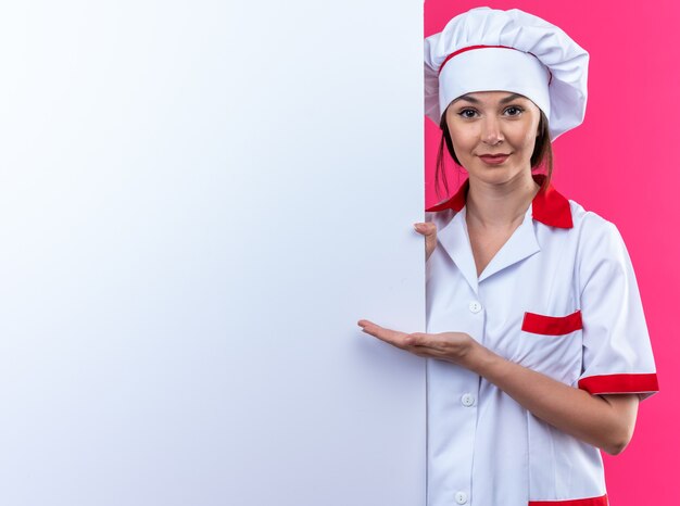 Feliz joven cocinera vistiendo uniforme de chef se encuentra y señala con la mano en la pared blanca cercana aislada en la pared rosa con espacio de copia