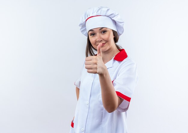 Feliz joven cocinera vistiendo uniforme de chef mostrando el pulgar hacia arriba aislado en la pared blanca