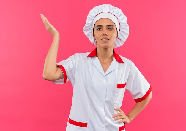 Feliz joven cocinera vistiendo uniforme de chef poniendo la mano en la cadera y levantando la mano sobre la pared rosa aislada con espacio de copia