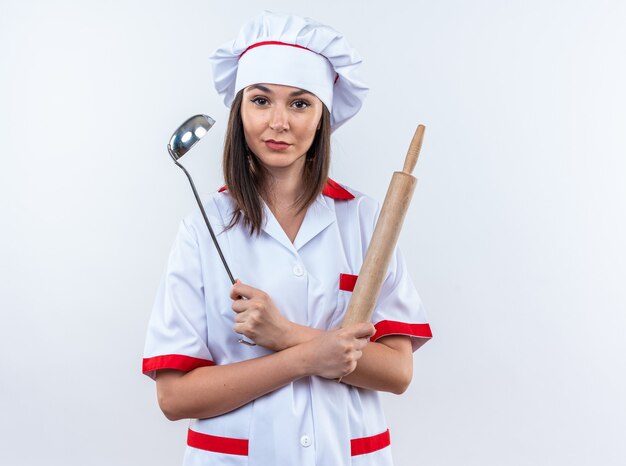 Feliz joven cocinera vistiendo uniforme de chef sosteniendo y cruzando la cuchara con rodillo aislado en la pared blanca