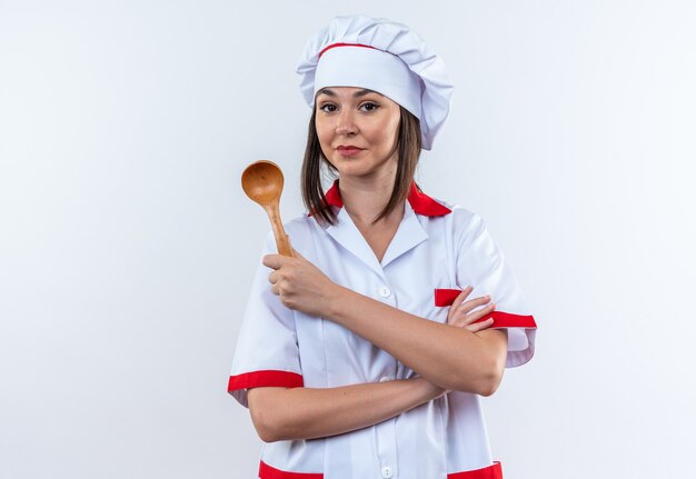 Feliz joven cocinera vistiendo uniforme de chef sosteniendo una cuchara cruzando las manos aislado en la pared blanca