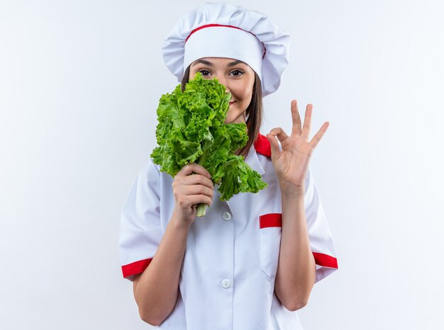 Feliz joven cocinera vistiendo uniforme de chef sosteniendo ensalada mostrando bien gesto aislado en la pared blanca