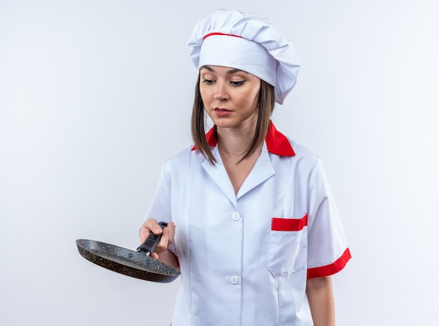 Foto gratuita feliz joven cocinera vistiendo uniforme de chef sosteniendo y mirando una sartén aislado en la pared blanca