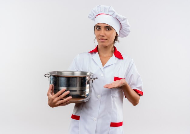 Feliz joven cocinera vistiendo uniforme de chef sosteniendo y puntos con una cacerola de mano en la pared blanca aislada con espacio de copia