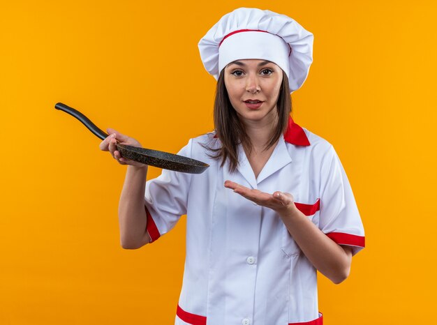 Feliz joven cocinera vistiendo uniforme de chef sosteniendo y puntos con la mano en la sartén aislado en la pared naranja