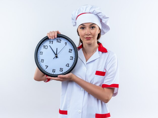 Feliz joven cocinera vistiendo uniforme de chef sosteniendo el reloj de pared aislado en la pared blanca