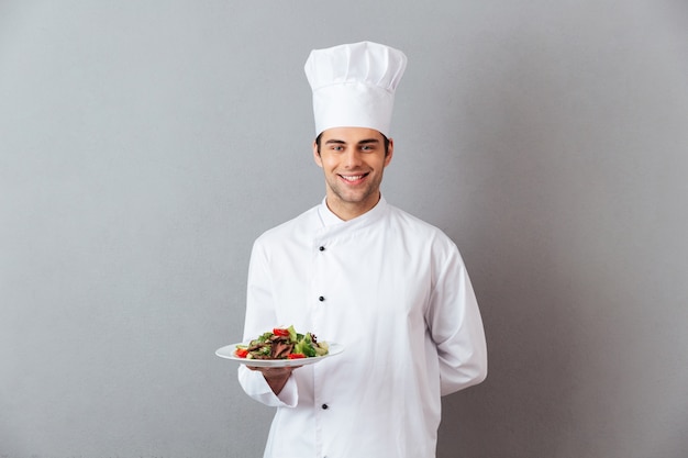 Feliz joven cocinero en uniforme con ensalada.