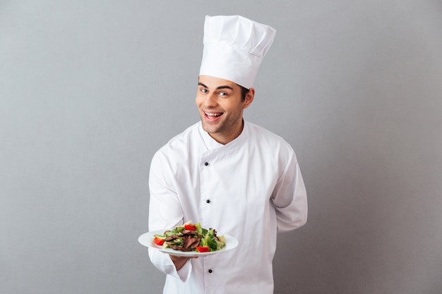 Feliz joven cocinero en uniforme con ensalada.