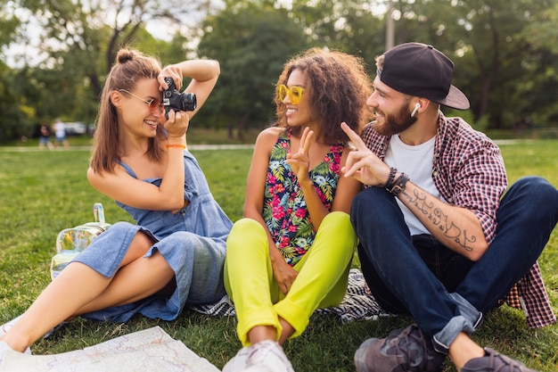 Feliz joven compañía de amigos sentados en el parque, hombres y mujeres divirtiéndose juntos, viajando con cámara, tomando fotografías