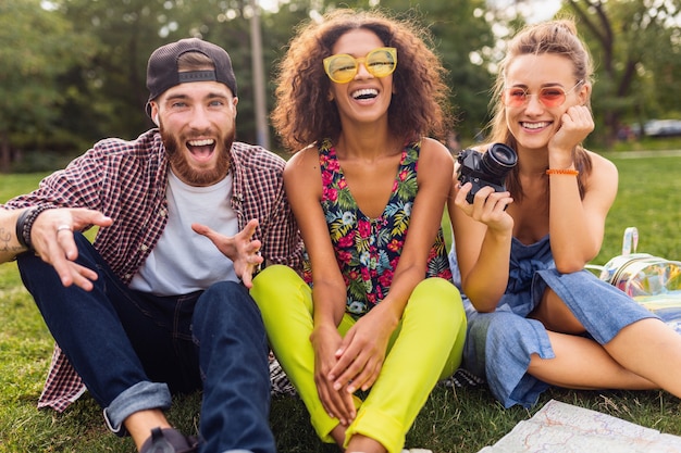 Foto gratuita feliz joven compañía de amigos sentados en el parque, hombres y mujeres divirtiéndose juntos, viajando con cámara,