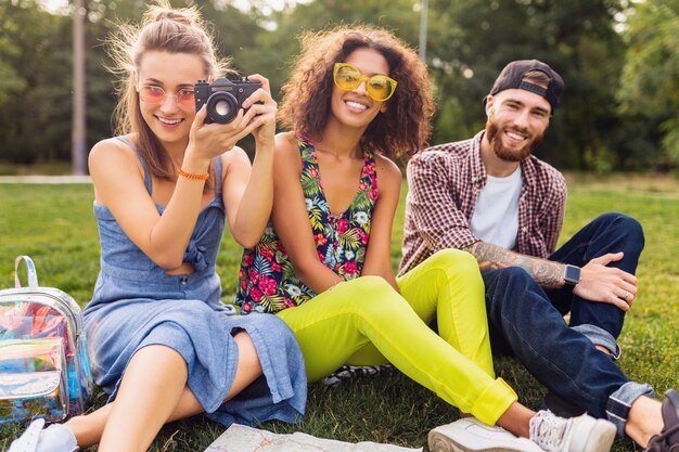Feliz joven compañía de amigos sentados en el parque, hombres y mujeres divirtiéndose juntos, viajando tomando fotos en la cámara, hablando, sonriendo