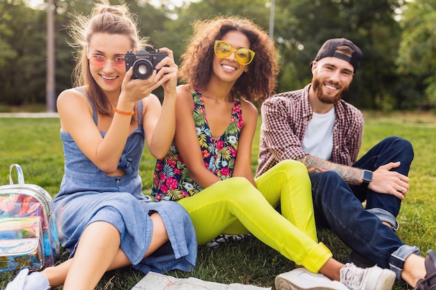 Feliz joven compañía de amigos sentados en el parque, hombres y mujeres divirtiéndose juntos, viajando tomando fotos en la cámara, hablando, sonriendo