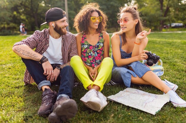 Feliz joven compañía de amigos sentados en el parque viajando mirando en el mapa de turismo, hombres y mujeres divirtiéndose juntos, estilo de moda hipster de verano colorido, tomando fotos en la cámara, hablando, sonriendo