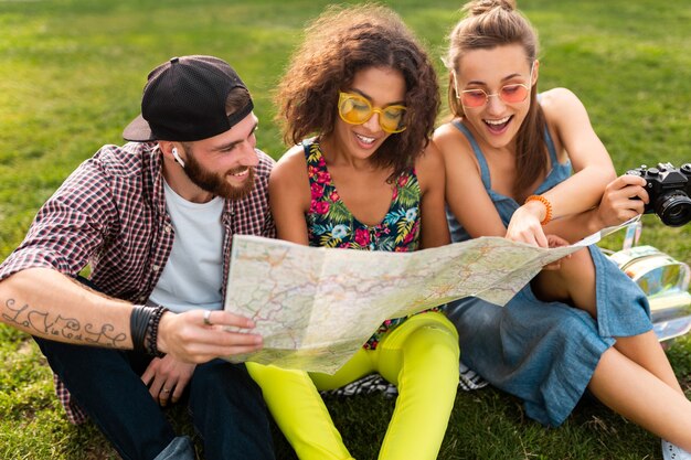 Feliz joven compañía de amigos sentados en el parque viajando mirando en el mapa de turismo, hombres y mujeres divirtiéndose juntos
