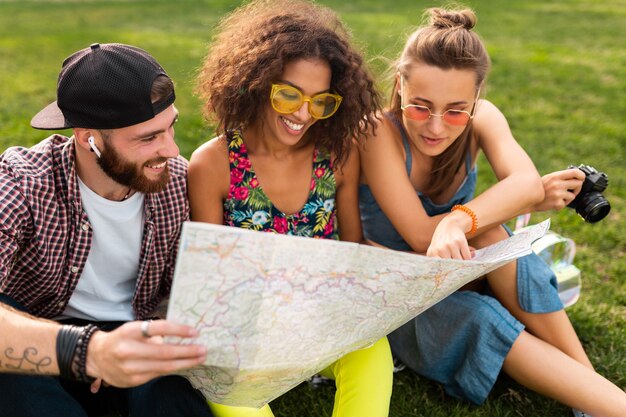 Feliz joven compañía de amigos sentados en el parque viajando mirando en el mapa de turismo, hombres y mujeres divirtiéndose juntos