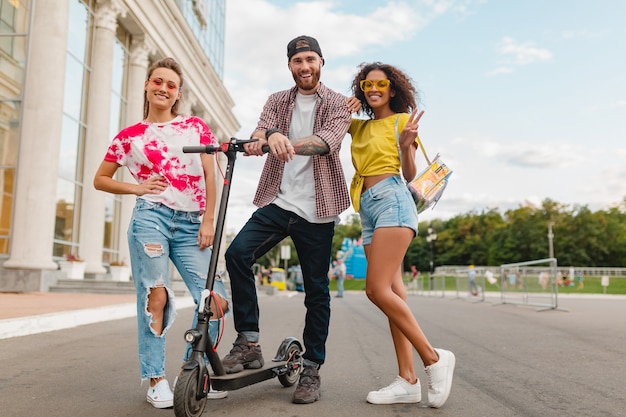 Feliz joven compañía de amigos sonrientes caminando en la calle con patinete eléctrico, hombre y mujer divirtiéndose juntos