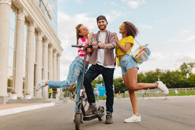 Feliz joven compañía de amigos sonrientes caminando en la calle con patinete eléctrico, hombre y mujer divirtiéndose juntos