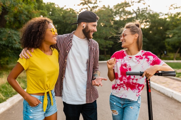 Feliz joven compañía de amigos sonrientes caminando en el parque con patinete eléctrico, hombre y mujer divirtiéndose juntos