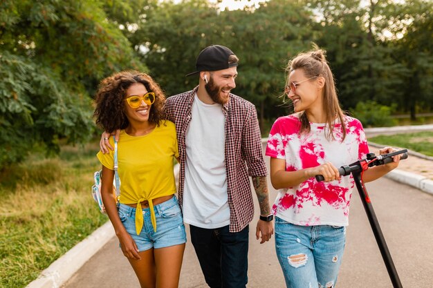 Feliz joven compañía de amigos sonrientes caminando en el parque con patinete eléctrico, hombre y mujer divirtiéndose juntos