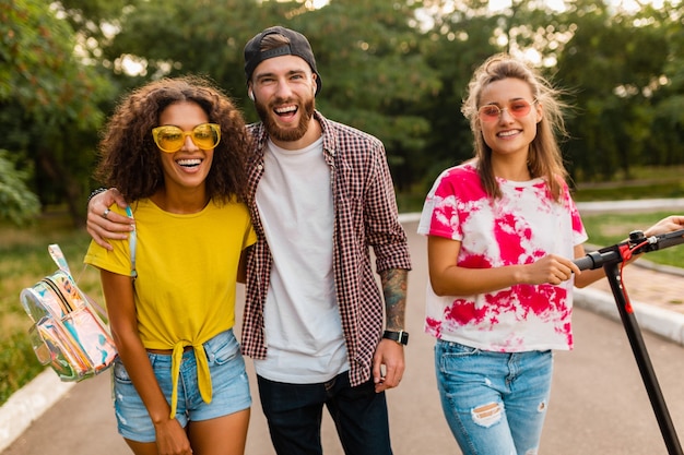 Feliz joven compañía de amigos sonrientes caminando en el parque con patinete eléctrico, hombre y mujer divirtiéndose juntos