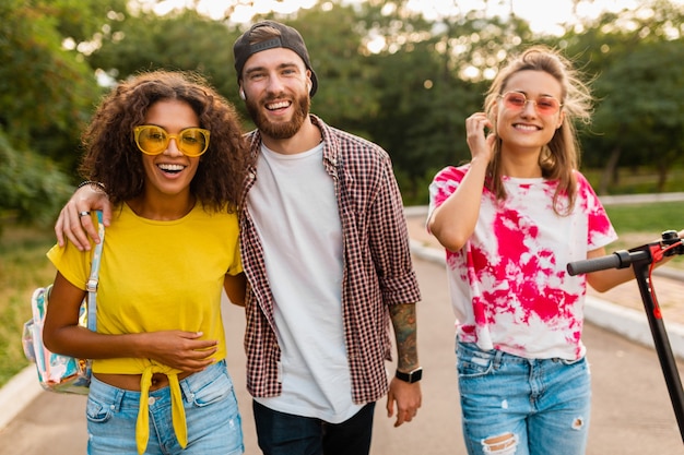 Feliz joven compañía de amigos sonrientes caminando en el parque con patinete eléctrico, hombre y mujer divirtiéndose juntos