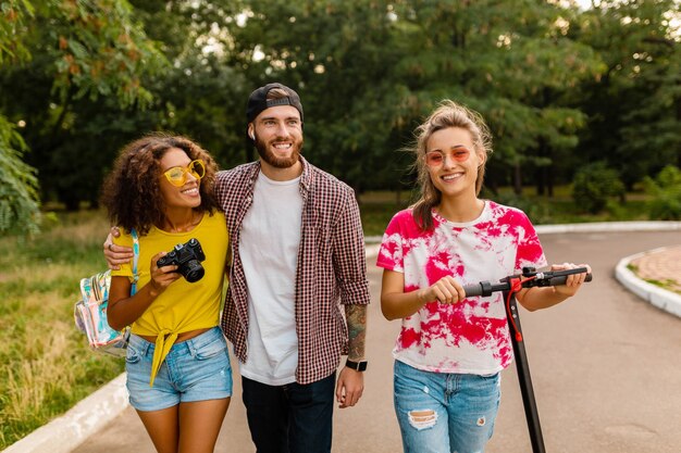 Feliz joven compañía de amigos sonrientes caminando en el parque con patinete eléctrico, hombre y mujer divirtiéndose juntos