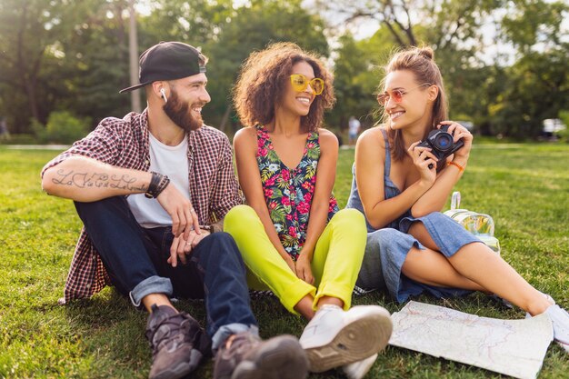 Feliz joven compañía de amigos sonrientes hablando sentado en el parque, hombres y mujeres divirtiéndose juntos, estilo de moda hipster de verano colorido, viajando con cámara