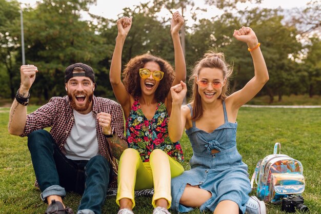 Feliz joven compañía de amigos sonrientes sentado en el parque sobre el césped, hombre y mujer divirtiéndose juntos