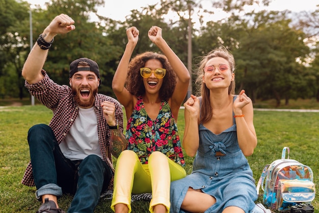 Foto gratuita feliz joven compañía de amigos sonrientes sentado en el parque sobre el césped, hombre y mujer divirtiéndose juntos