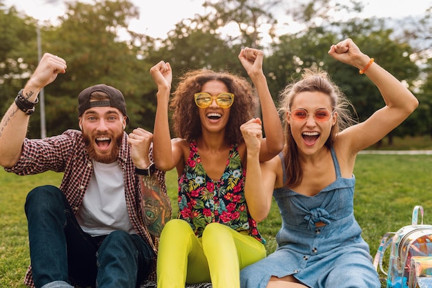 Feliz joven compañía de amigos sonrientes sentado en el parque sobre el césped, hombre y mujer divirtiéndose juntos