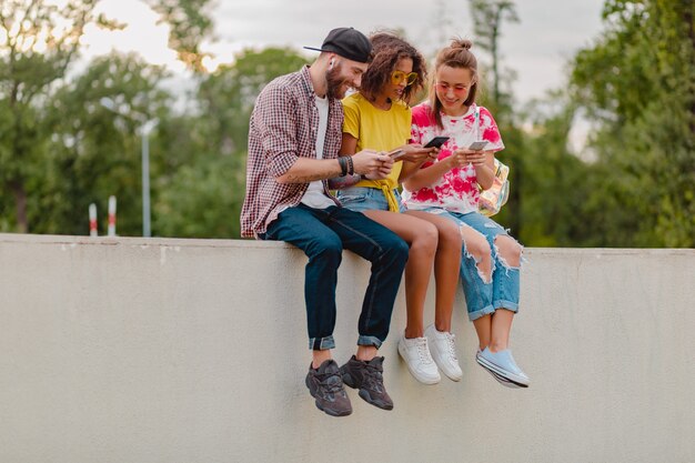 Feliz joven compañía de amigos sonrientes sentado parque usando teléfonos inteligentes