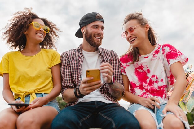 Feliz joven compañía de amigos sonrientes sentado parque usando teléfonos inteligentes