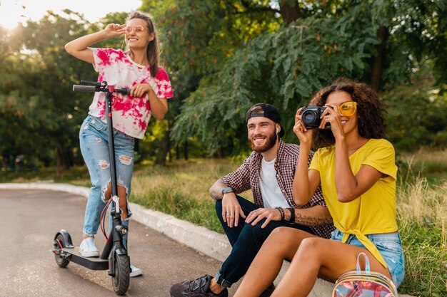 Feliz joven compañía de amigos sonrientes sentados en el parque sobre el césped con patinete eléctrico, hombre y mujer divirtiéndose juntos