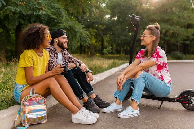 Feliz joven compañía de amigos sonrientes sentados en el parque sobre el césped con patinete eléctrico, hombre y mujer divirtiéndose juntos