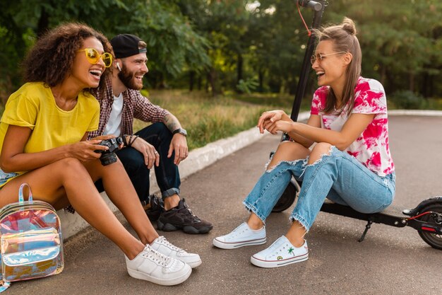 Feliz joven compañía de amigos sonrientes sentados en el parque sobre el césped con patinete eléctrico, hombre y mujer divirtiéndose juntos